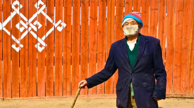 Elderly man in Ulaanbaatar wearing a mask and with walking stick in front of a wooden fence