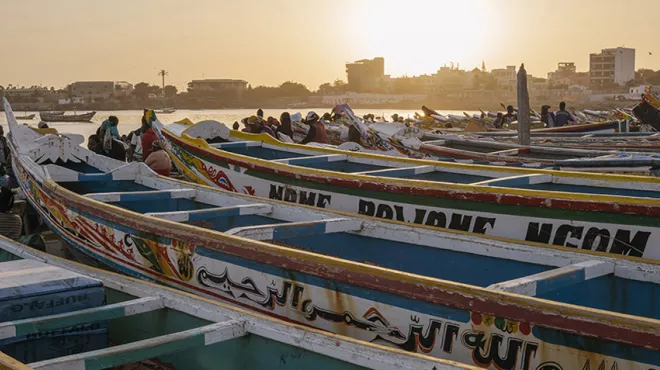 Several Pirogues lying at the shore of Dakar with people sitting in between and with city skyline in fading sunlight in the background