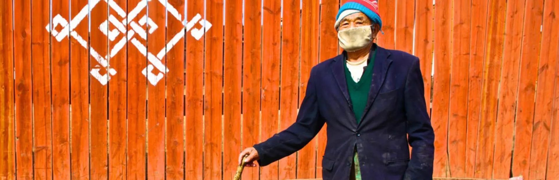 Elderly man in Ulaanbaatar wearing a mask and with walking stick in front of a wooden fence