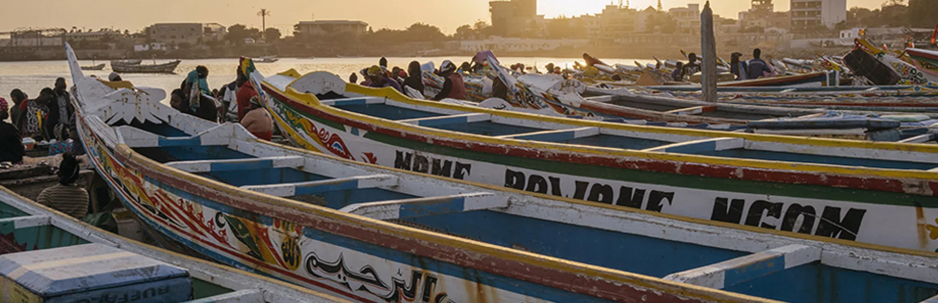 Several Pirogues lying at the shore of Dakar with people sitting in between and with city skyline in fading sunlight in the background